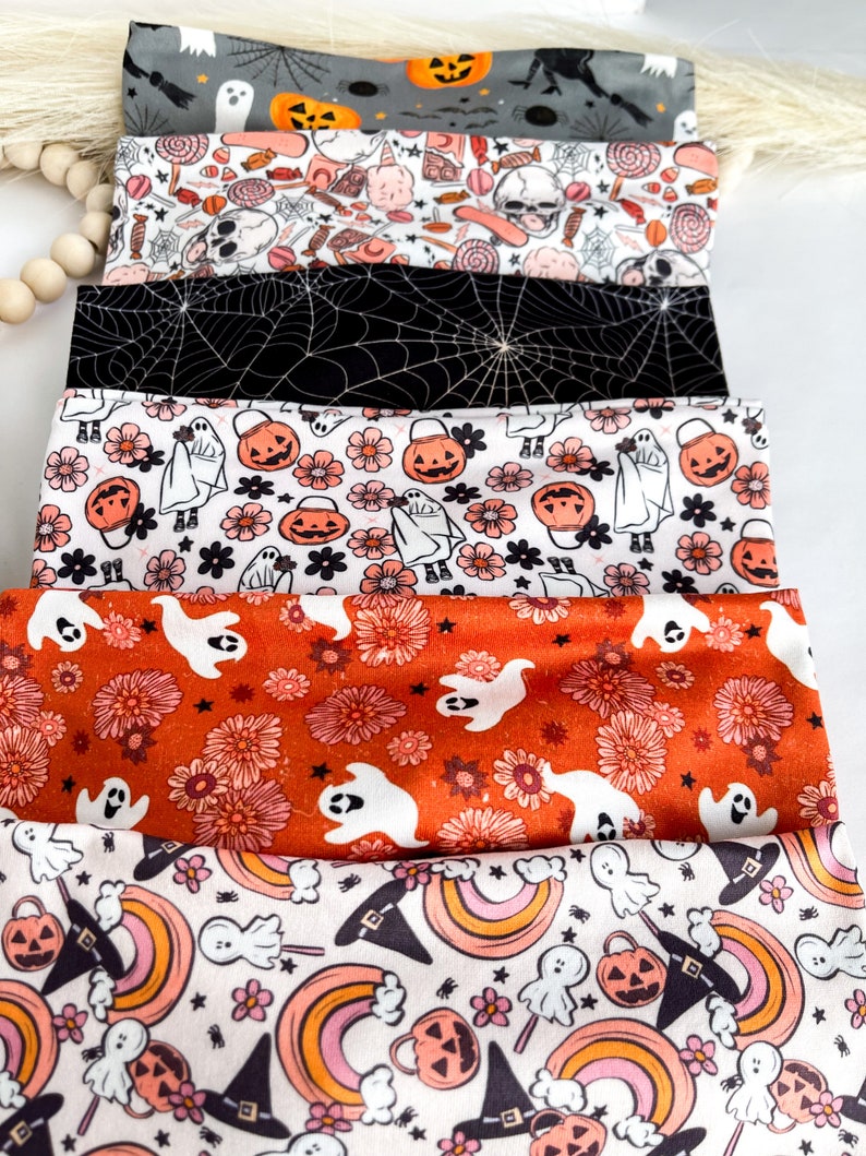 Może przedstawiać: Stos materiałowych opasek do włos&oacute;w z motywem Halloween. Opaski mają r&oacute;żne wzory, w tym duchy, dynie, pajęczyny i kwiatowe wzory. Kolory to czarny, biały, pomarańczowy i szary. Opaski prawdopodobnie na uroczystą okazję.