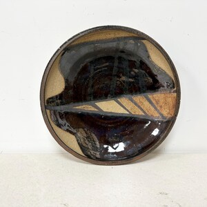 May include: A handmade ceramic plate with a leaf design. The plate features a dark brown, glossy center with a tan and brown leaf pattern. The rim is a lighter tan color.