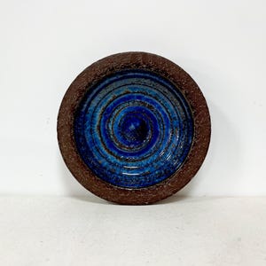 Może przedstawiać: Okrągła, ceramiczna miska z teksturowanym, ciemnobrązowym brzegiem i żywym niebieskim wzorem spirali pośrodku. Wnętrze miski zdobią koncentryczne okręgi w różnych odcieniach niebieskiego, tworząc atrakcyjny wizualnie wzór.