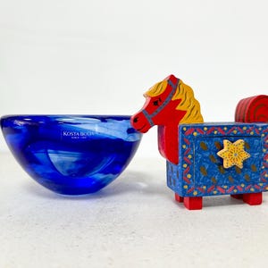 May include: A blue glass bowl with white swirls and the text "KOSTA BODA SINCE 1742" next to a colorful wooden horse figurine. The horse is red, blue, yellow, and has a star on its side.