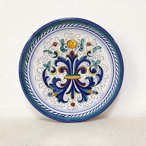 Può includere: Un piatto rotondo in ceramica con un design dipinto a mano che presenta un motivo centrale a fleur-de-lis blu, accentuato da motivi floreali gialli, verdi e marroni. Il bordo è decorato con un motivo a corda intrecciata blu e bianca e strisce verdi.