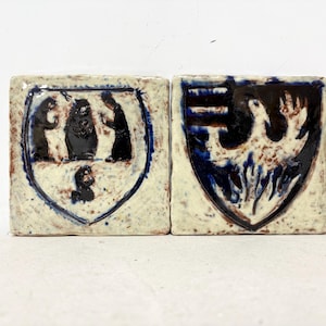May include: Two square ceramic tiles with hand-painted heraldic designs. One tile features a blue shield with black figures, and the other depicts a phoenix in blue and brown. The tiles have a textured, off-white background.
