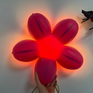 May include: A pink flower-shaped night light with a red LED center. The light is mounted on a white wall.