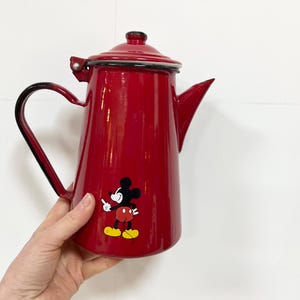 May include: A red enamel coffee pot with a black handle and spout. The pot has a Mickey Mouse decal. The pot is held by a hand. The coffee pot is approximately 20 cm tall.