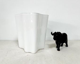 May include: A white, wavy-edged vase stands next to a small, black figurine of an elephant. The vase has a glossy finish and a unique, organic shape. The elephant figurine is detailed and has a smooth texture.