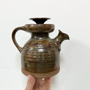 May include: A handmade ceramic teapot with a lid. The teapot is a mix of brown, green, and yellow hues, with a textured surface. It features a curved handle and a spout. The base is a lighter brown colour.