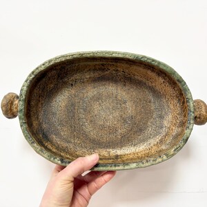 May include: Oval ceramic serving dish with a textured, speckled brown interior and a green-tinged rim. The dish has two small, rounded handles. The handmade pottery has a rustic, earthy aesthetic, suitable for serving food.