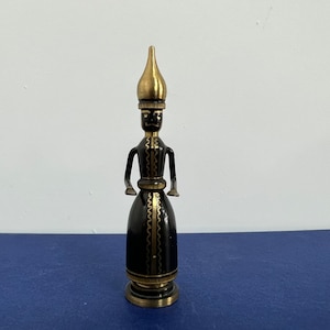 May include: A black and gold metal figurine of a person wearing a tall pointed hat. The figurine is standing on a base with a decorative design.