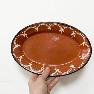 May include: Oval terracotta serving platter with a black rim and white scalloped floral design. The interior is a solid reddish-brown color. A decorative piece for serving food or displaying items.