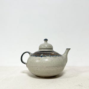 May include: A ceramic teapot with a speckled, off-white glaze. The teapot features a dark brown and blue band around the middle and a matching lid. The handle and spout are dark blue. A classic piece for serving tea.