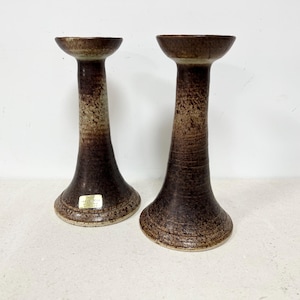 May include: Two ceramic candle holders with a textured, speckled brown and tan finish. The candle holders have a flared base, a long, slender stem, and a small, flat top. The candle holders are approximately 8 inches tall.