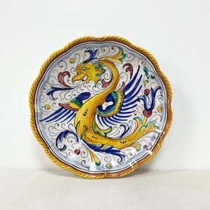 May include: Ceramic decorative plate with a scalloped edge. The plate features a yellow, blue, and green mythical creature design on a white background. The rim is a textured yellow and brown.