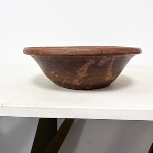 May include: A rustic, brown earthenware bowl with a wide, shallow design. The bowl has a rounded rim and a slightly textured surface. The exterior features lighter brown markings, adding to its aged appearance. The bowl sits on a white surface.