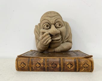 May include: A tan-colored stone sculpture of a face with a large nose and hands near the face, resting on a brown leather-bound book. The book has decorative embossed patterns.