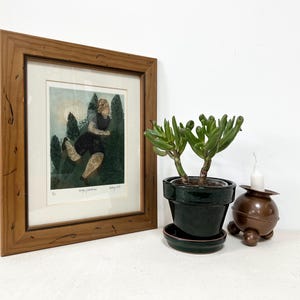 Może przedstawiać: Oprawione dzieło sztuki z brązową drewnianą ramą. Grafika przedstawia osobę biegnącą przez las. Obok ramy znajduje się zielona roślina doniczkowa i brązowy świecznik.