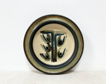 May include: A round ceramic plate with a cream-colored center and a dark blue and brown rim. The center features a stylized tree design in blue and brown. The plate is approximately 8 inches in diameter.