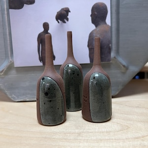 May include: Three handcrafted ceramic bottles. Each bottle has a reddish-brown base and a speckled, grey-green oval design. The bottles are displayed on a wooden surface. The bottles are likely handmade.