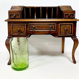 May include: A dark wood writing desk with a carved design and multiple drawers. The desk has a raised back with a series of open compartments. The desk is sitting on a white surface with a green glass jar in front of it.