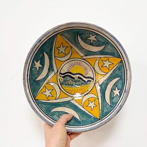 Pode incluir: Uma tigela de cerâmica redonda com um design circular central de um sol amarelo sobre ondas azuis e peixes pequenos. Uma grande estrela amarela de cinco pontas sobrepõe o fundo, rodeada por luas crescentes brancas e estrelas em fundo turquesa.