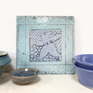 Pode incluir: Um azulejo de cerâmica quadrado com acabamento esmaltado azul claro, apresentando um desenho de linhas azul-escuras de uma figura com os braços levantados sobre um grupo de círculos. O azulejo é emoldurado por triângulos.