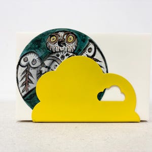 May include: A decorative napkin holder featuring a hand-painted owl design on a teal background. The holder is paired with a bright yellow cloud-shaped napkin holder, adding a pop of color and whimsy to any table setting.
