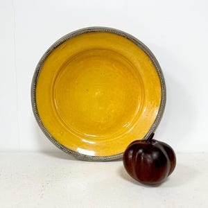 Può includere: Un piatto di ceramica giallo con bordo grigio e una piccola zucca marrone scuro. Il piatto ha un design ampio e poco profondo. La zucca è rotonda con una superficie strutturata. Gli oggetti sono su una superficie bianca.