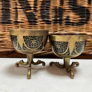 May include: Two gold-coloured egg-shaped cups with black and gold decorative designs and three legs each.