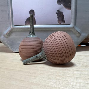 Può includere: Due tappi per bottiglia in ceramica fatti a mano, color terracotta. Ogni tappo ha un corpo rotondo con linee incise e un manico sottile e maculato. Un tappo è in posizione verticale, l'altro è appoggiato su un lato. Lo sfondo include un'immagine incorniciata e una superficie in legno.