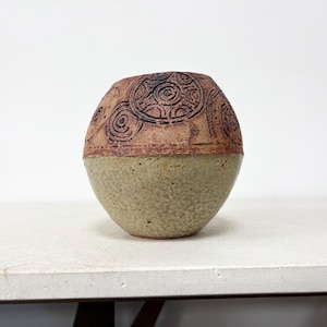 May include: A round ceramic vase with a two-tone finish. The upper portion features a textured reddish-brown surface with carved circular spiral patterns, while the lower portion has a speckled, light beige matte finish with a rough texture.