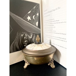 May include: A brass trinket box with a mother of pearl lid and a decorative finial. The box is sitting on a book with the text "A Foreword:  Master of the Wonderful Things He Does" visible.