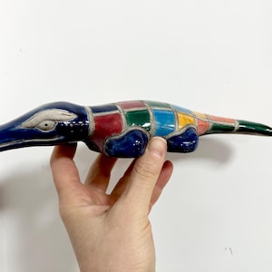 May include: A ceramic alligator figurine with a patchwork design. The alligator's head and feet are dark blue, with a white eye. The body is a mosaic of red, green, yellow, and blue squares. The tail is green.