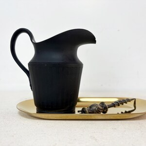 May include: A matte black pitcher with a curved handle and spout sits on a gold-colored oval tray. A vintage corkscrew rests on the tray. The items are displayed against a white background.
