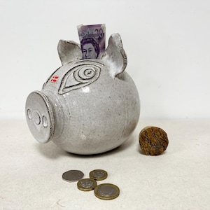 May include: A ceramic piggy bank in the shape of a pig, with a slot for inserting money. The bank is light gray with black detailing and a small red square. A twenty-dollar bill is visible in the slot, with coins and a cork nearby.