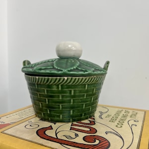 May include: A green ceramic butter dish with a lid shaped like a basket with a white knob. The dish is sitting on a yellow book with the text "The Regional Cooking of Italy" printed on the cover.