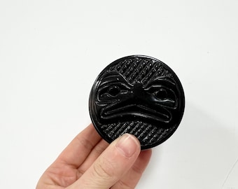 Può includere: Un oggetto decorativo rotondo e nero con un disegno di volto scolpito. L'oggetto ha una superficie strutturata ed è tenuto in mano su uno sfondo bianco. Il volto sembra essere un uccello o un animale stilizzato.