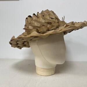 May include: A woven straw hat with a wide brim, displayed on a mannequin head. The hat is made of light brown, dried plant material, with a textured, layered design. The hat's brim is approximately 16 inches in diameter.