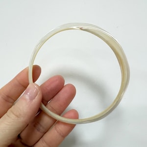 Op de afbeelding: Een cirkelvormige armband met een glad, gepolijst oppervlak. Het materiaal heeft een doorschijnende, gebroken witte uitstraling met subtiele, wervelende parelmoertinten die het licht vangen en een zachte, iriserende glans over de band creëren.