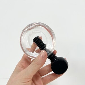 May include: A clear glass bottle stopper with a black screw and a round black top. The stopper is designed to fit into a bottle opening. The image is set against a white background.