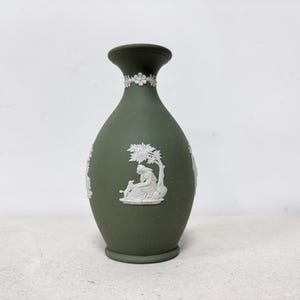 Può includere: Un vaso in ceramica verde scuro con figure in rilievo bianche e dettagli floreali. Il vaso ha un corpo arrotondato, un collo stretto e un bordo svasato. Il rilievo bianco raffigura figure e scene classiche.