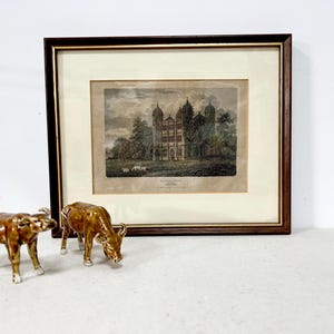 May include: An antique framed print of a stately building with a detailed facade, set in a landscape with trees and sheep. Two ceramic bull figurines with brown and gold glaze are in the foreground.