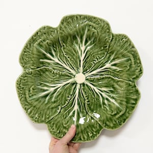 May include: A green ceramic cabbage-shaped serving platter. The platter features a detailed leaf design with white veins and a cream-coloured centre. The edges of the platter are scalloped, and the overall shape resembles a large cabbage.