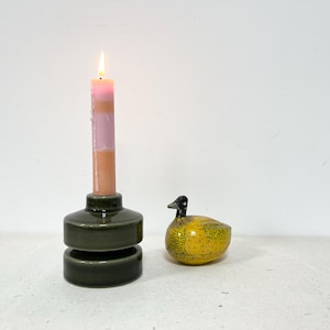 May include: A burning candle with pink and tan stripes sits in a dark green ceramic candle holder. A yellow duck ornament with black details is next to the candle holder. The items are on a white surface.
