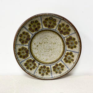 May include: A circular ceramic plate with a brown edge and a cream-coloured centre. It features a repeating floral pattern in brown and olive green, with blue lines. The plate has a rustic, handmade appearance, typical of pottery.