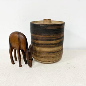 May include: A wooden deer figurine with a dark brown finish, positioned near a cylindrical ceramic container. The container has a lid and is primarily brown with dark brown horizontal stripes. The items are on a white surface.