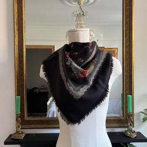May include: A black square scarf with a floral pattern in red, green, and white. The scarf has a fringed edge and a decorative border. The scarf is displayed on a mannequin in front of a gold-framed mirror.
