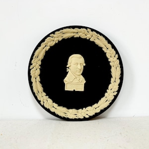 May include: A black and cream ceramic plate with a raised image of a man's bust in the center. The plate is decorated with a cream-colored wreath around the edge.