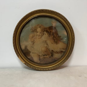 May include: A vintage, round framed print featuring two cherubic figures in soft, muted tones. The gold-toned frame has a decorative edge. The artwork depicts the children in a tender moment, with one whispering to the other.