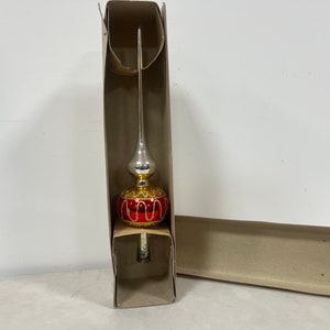 May include: A glass Christmas tree topper featuring a tall, slender silver spire above a round red bulb. The red bulb is decorated with gold glitter swirls and lines. The ornament is housed in a tall, narrow, light brown cardboard box.