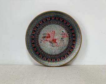 May include: A round ceramic plate with a gold rim and a black and red design. The design features a chariot pulled by two horses and a person driving the chariot.