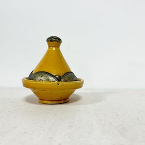 Puede incluir: Una olla de tagine de cerámica amarilla con tapa cónica y una banda metálica decorativa alrededor de la base. El tagine es un recipiente de cocina tradicional del norte de África, ideal para guisos y tagines a fuego lento.